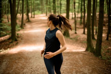 Ormanda ağaçların arasında koşan uzun saçlı, arkası ve önü manzaralı, sportif bir kadın. Fitness, vücut sağlığı ve ruhsal durum için koşma.