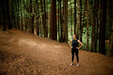 Spordan, egzersizden ya da eğitimden sonra dinlenen genç atletik kadının yatay çekimleri. Rahat koşucu, güzel orman arka planına karşı duruyor.