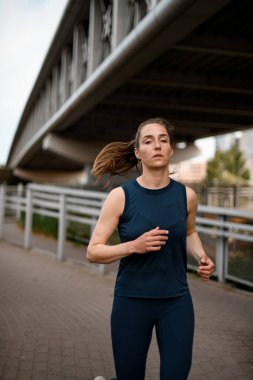 Modern köprünün yanındaki caddede spor kıyafetleriyle koşan genç bir kadın. Spor, sağlık, spor, kilo kaybı, kardiyo eğitimi kavramı