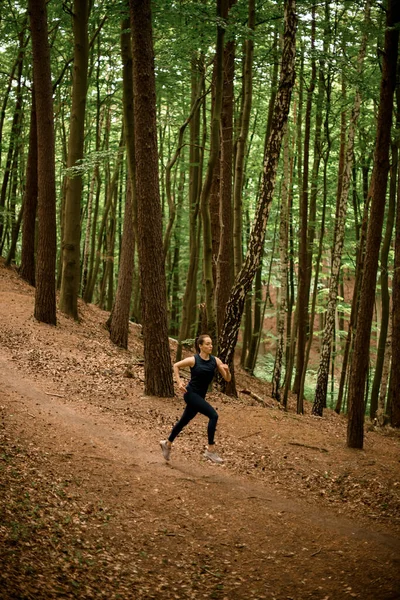 Athlete young woman in black sportswear jogging downhill on forest trail, tall leafy trees on background. Fit girl jogging outside. Healthy lifestyle. Sporty and activity concept