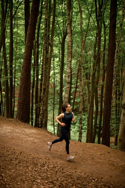 Young fit woman in black sportswear jogging downhill on forest trail, tall leafy trees on background. Slim girl jogging outside. Healthy lifestyle. Sporty and activity concept