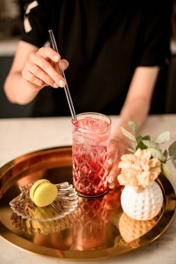 Müşteri siparişi veren metal tepsi: limonata, tatlı tatlı makaron, kafenin içinde çiçekler. Garson kız bardağa limonatayla saman sokuyor.