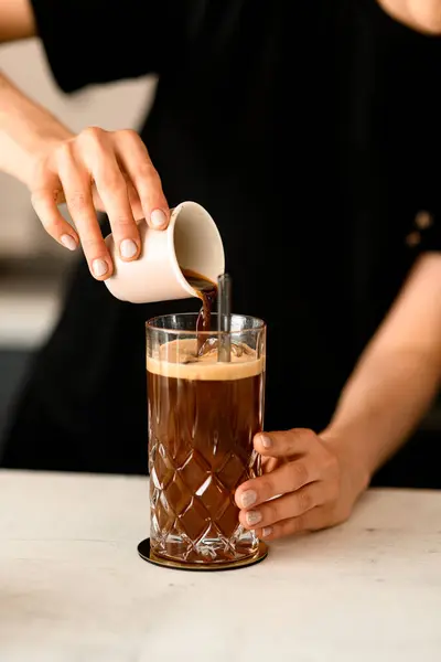 Female barista pouring coffee on ice cubess in high old fashioned glass with tonic served on table in coffee shop or restaraunt. Close up view. Coffee break.