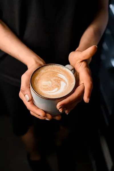 Woman holding in hands cup with tasty steaming cappucino decorated with foamy art, top view. Hot served coffee in womans hands