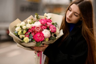 Siyah paltolu genç bir kadın elinde protea, gül ve el işi kağıdına sarılmış şakayık demetleriyle büyük bir çiçek buketi tutuyor. Kız açık havada çiçek karışımı bir buket tutuyor. Çiçekçi dükkanı konsepti