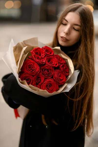 Beautiful brunette woman with large bouquet of red roses for holiday. Attractive woman with flowers bouquet standing on street. Close-up portrait. Blurred background