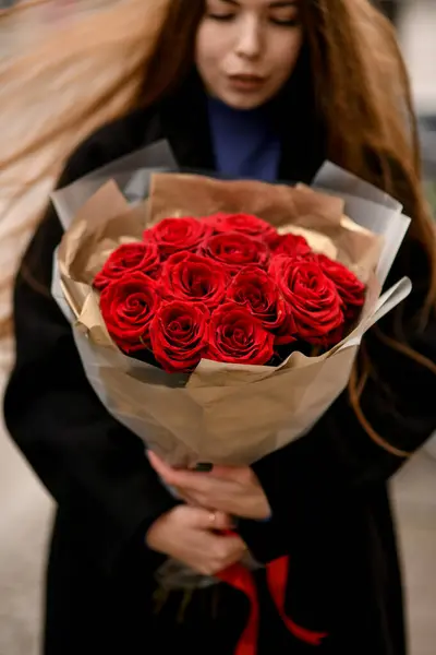 Beautiful view on large bouquet with red roses in brown wrapping paper in woman brunette hands. Young woman holding bouquet of roses flowers on the street