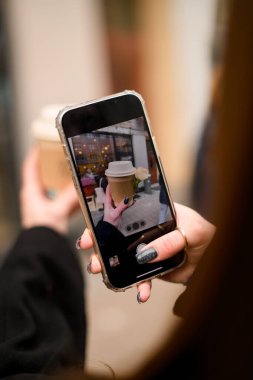 Mobil ekranın seçici odağı, kadın elinde gül çiçeğiyle süslenmiş kağıt bardaktan yapılmış kahve fotografı. Kadın eli, kağıt kahve fincanının fotoğrafını çekiyor.