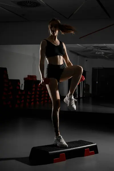 带马尾辫的女健身教练用哑铃进行体育锻炼 站在体操台上背对着镜子 体操和生活方式概念 — 图库照片