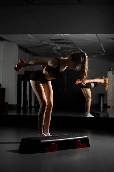 全神贯注的年轻女子用哑铃进行体育锻炼 站在黑暗体育馆的台阶台上 背对着镜子 培训和健康生活方式概念 — 图库照片