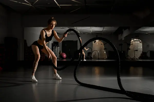 stock image Young fit sportswoman working out in functional training gym doing crossfit exercise with battle ropes against mirror. Battle ropes session. Concept: power, strength, healthy lifestyle, sport.