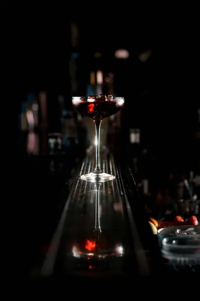 View on tasty red cocktail in high martini glass with ice cube reflected in steel surface of bar counter. Dark blurred background. Menu of classic cocktails