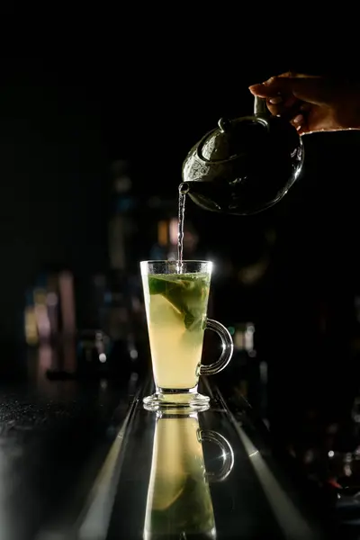 Female barmans hand pours hot liquid from beautiful clay teapot into transparent high glass on blurred bar counter background. Close up. Space for text