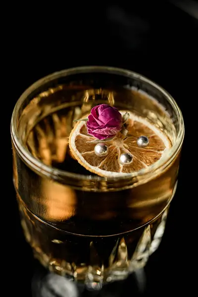 Whiskey glass with transparent yellow cocktail decorated with dried orange, silver beads and little rose flower, front view. Dark background