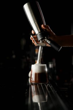 Çelik sifonlu barmen kız bardağa soğuk kahve ile beyaz köpük ekler. Karanlık arka plan. Seçici odak