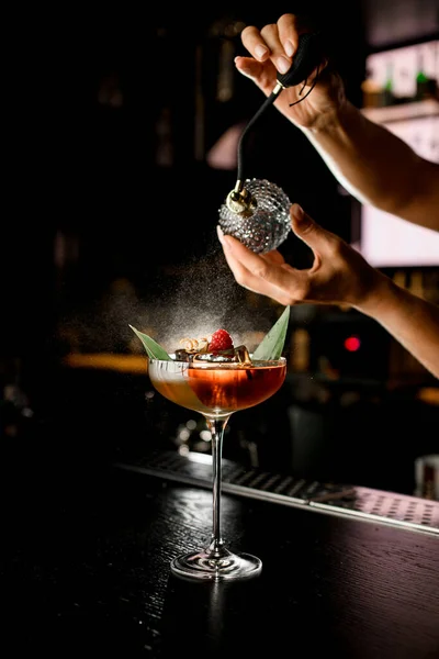 Female barman hand spraying transparent bitter on martini glass with elegant red cocktail decorated with berries and banana leaf on bar counter. Bartender finishes making the cocktail at bar counter