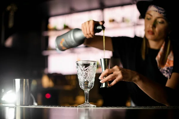 View of girl bartender at bar counter holding bottle with syrup and pours it to steel jigger for making cocktail. Woman bartender creates tasty alcoholic drink