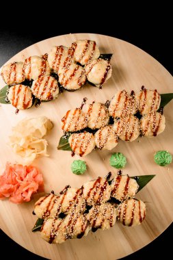 Siyah arka planda koyu unagi soslu yuvarlak ahşap bir tahtanın üzerinde yılan balıklı suşi ruloları. Beyaz ve kırmızı kızıl, wasabi.