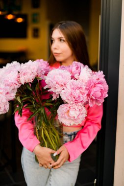 Parlak pembe gömlekli güzel esmer kız açık pembe şakayıklı cam bir vazo tutuyor.