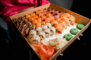 Pembe bluzlu kadın elleri, ellerinde bir karton kahverengi kutuda wasabi, kırmızı ve beyaz zencefilli suşi ruloları tutuyor.