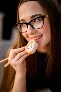 Gözlüklü uzun saçlı, tatlı, sırıtan esmer kadın yemek çubuklarıyla tutulmuş suşi rulolarının tadını çıkarıyor.