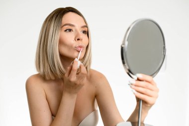 Çıplak omuzlu güzel bir kadın yuvarlak aynanın önünde dudaklarını parlatıyor.