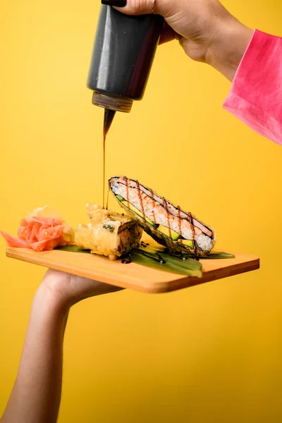 Female hand holds a tray with two halves of a sushi sandwich and red ginger on an orange background. Another hand pours sauce from a black bottle over the sushi