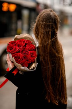 Siyah ceketli güzel bir kadın kameraya arkasını döndü ve bulanık bir caddenin arka planında lüks güller taşıyan bir rezervatörü tuttu.