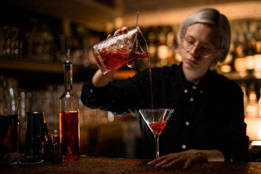 Bardaklı sarışın kadın barmen bardaktan bardağa hazır bir kokteyl dolduruyor bulanık bir tezgahta.