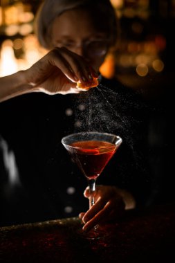 Yoğunlaşmış kadın barmen portakal dilimini sıkıyor ve bulanık arka planda kırmızı bardakta portakal kabuğu serpiyor.