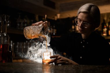 Barmen bardaktan dumana karışan bir kokteyli bardağın arka planında sadece buz olan bir bardağa doldurmaya başlar.