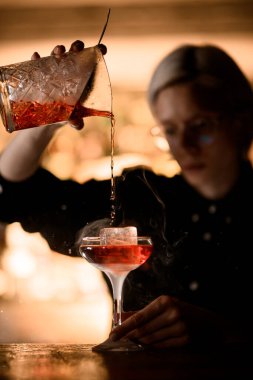 Kadın barmen dumanlı kokteyl yapmaktan zevk alıyor. Karıştırma bardağından buzlu bardağa döküyor.