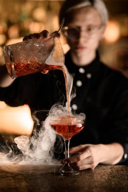 Özenli bayan barmen bardakla süzgeci karıştırıp büyük buz parçasıyla karıştırarak füme kokteyl dolduruyor.