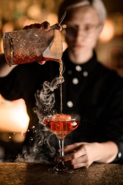 Özenli bayan barmen bardakla süzgecin karışımından tütsülenmiş kokteyl dökerek büyük bir buz parçasıyla farklı yönlere sıçrıyor.