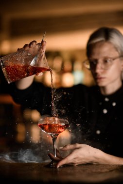 Kadın barmen yavaşça karıştırıcı bardaktan dumanlı bir kokteyl dökerek büyük bir buz parçasıyla bardağa döküyor.