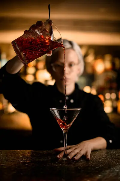 Ready red cocktail pouring into a cocktail glass on a blurred background of a female bartender and a bar counter