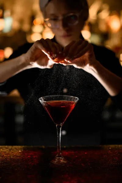 Serious blond woman bartender in glasses splashing freshly squeezed orange peel into ready red cocktail in cocktail glass