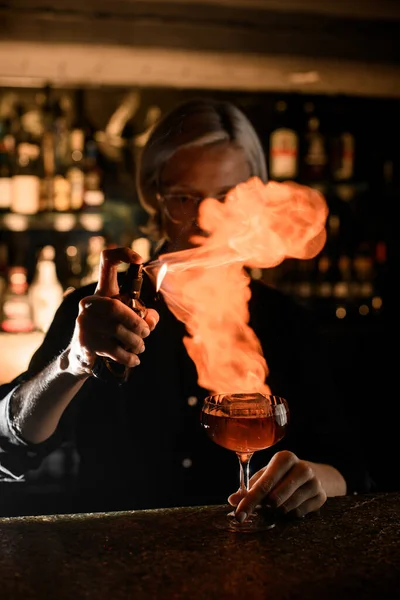 Female bartender makes fire cocktail with spray and lighted match on bar counter against background of bar showcase