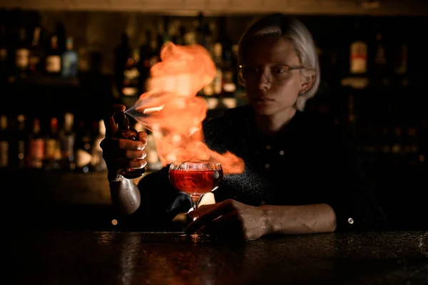 Female bartender carefully prepares fire cocktail with spray and lighted match on bar counter against background of bar showcase