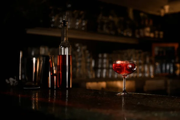 Stemmed glass with a cocktail stands on the bar next to bottles and other bartender accessories on the background of a bar showcase