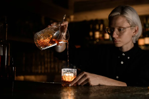 Female bartender pours a cocktail from a high-raised mixing glass into another glass with ice on the background of a bar showcase