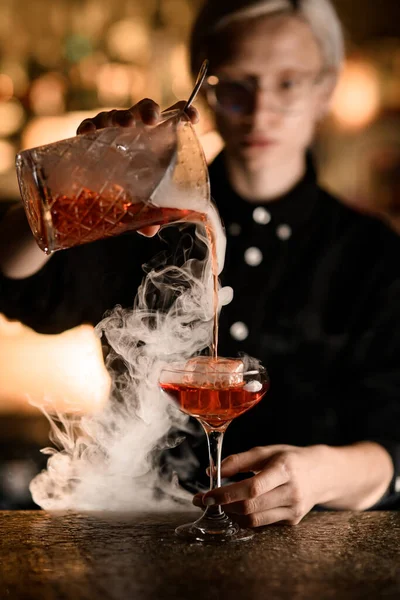 Female bartender enjoying the process of pouring a cocktail from a mixing glass into a glass with ice. Image of the bartender is blurred in the background