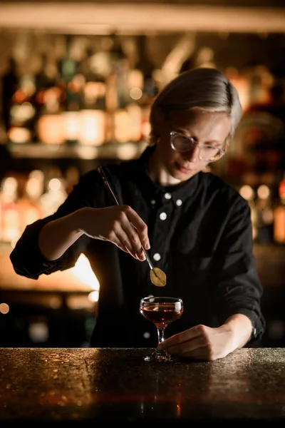 Female blonde bartender in glasses adds a green leaf with tweezers to a brown cocktail in a stemmed glass on the bar counter