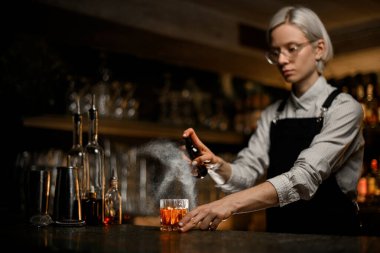 Açık renk üniformalı kadın barmen ve siyah önlüklü kadın arka planda sprey bulunan bardağa hazır bir kokteyl püskürtüyor.