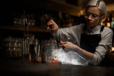 Kadın barmen alkollü içecekleri bir bardağa karıştırıyor, aynı anda barın yanındaki gayzer şişesinden döküyor, yakında duman görünüyor.