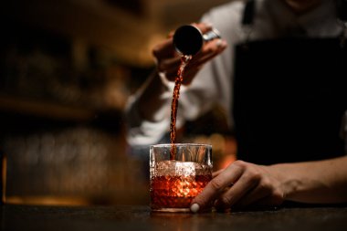 Beyaz manikürlü kadın eller kokteyl yapmak için bardağa alkollü içecek dökerler. Bulanık bir arka planda barın üstünde jigger tutarlar.