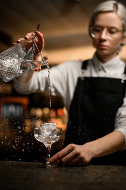 Bardaklı kadın barmen, büyük bir buz kütlesi olan saplı bir bardağa temiz bir içki dolduruyor, farklı yönlere doğru sıçrıyor.