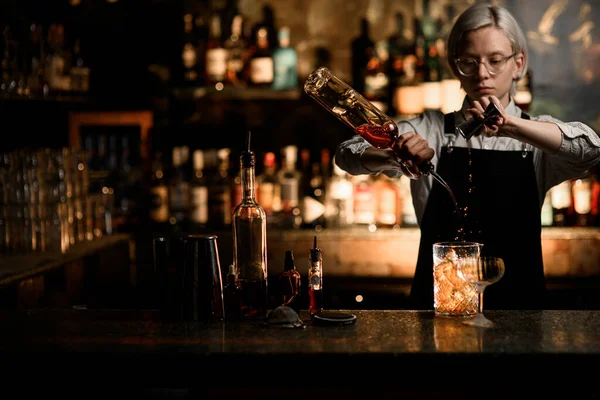 Female bartender pours an alcoholic drink into a mixing glass from a jigger and a bottle with a geyser, there is a bartenders equipment on the bar counter