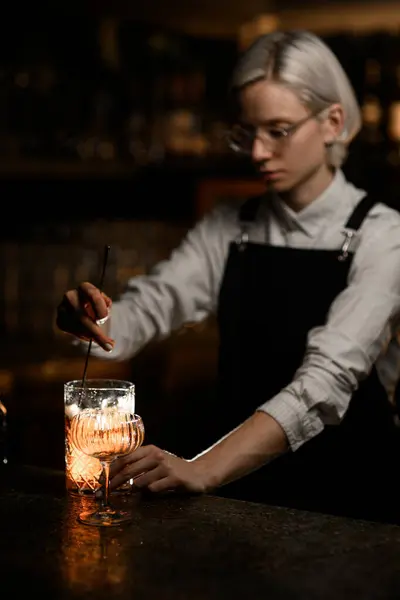 Female bartender in a light uniform with a black apron mixes a cocktail with ice in a mixing glass with a long bar spoon, side view