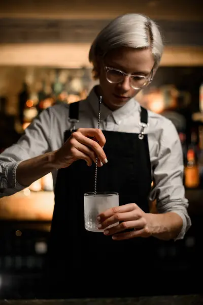 Female bartender stirs something in a frosted glass, which she holds in her hand, with a long bar spoon